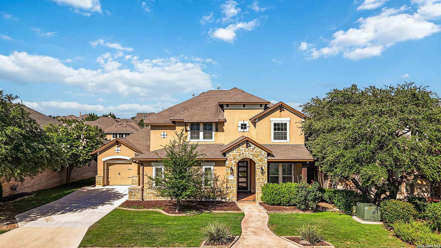 The Texas Hill Country front elevation of 28722 Front Gate in midday light, brown roof and stucco facade with side-facing garage entrance and mature trees flanking the front yard